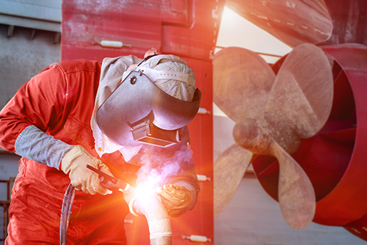 A shipyard worker welding metal beside a large ship propeller.