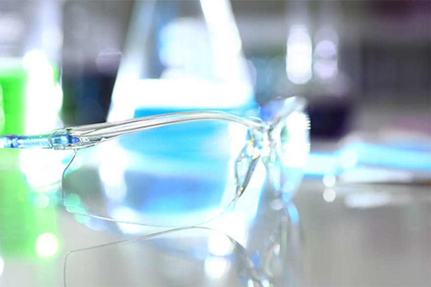 Clear protective safety glasses resting on a reflective pharmaceutical laboratory bench, with blurred lab glassware in the background.
